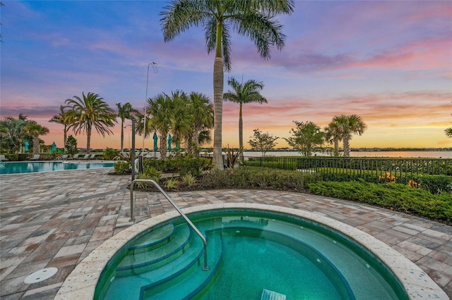 pool at dusk featuring a community hot tub, a water view, and a patio area