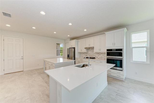 kitchen with white cabinets, appliances with stainless steel finishes, sink, an island with sink, and decorative backsplash