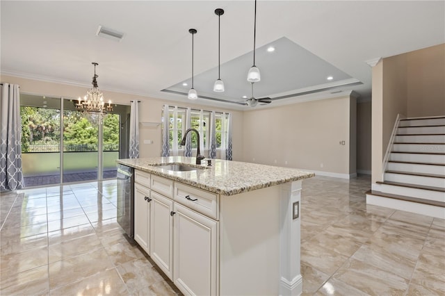 kitchen with ceiling fan with notable chandelier, light stone countertops, a tray ceiling, a kitchen island with sink, and sink