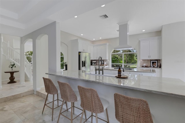 kitchen featuring light stone counters, white cabinetry, stainless steel fridge with ice dispenser, decorative backsplash, and island range hood