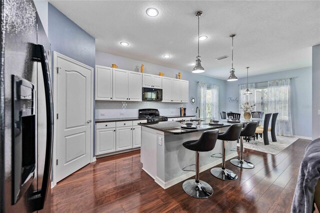 kitchen featuring black appliances, pendant lighting, an island with sink, dark wood-type flooring, and white cabinetry