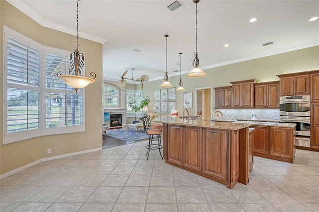 kitchen featuring stainless steel double oven, hanging light fixtures, crown molding, and a kitchen island with sink