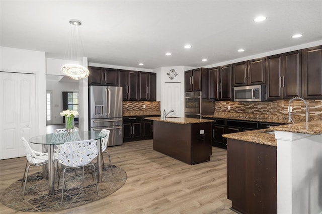 kitchen with decorative light fixtures, appliances with stainless steel finishes, an island with sink, light wood-type flooring, and stone counters