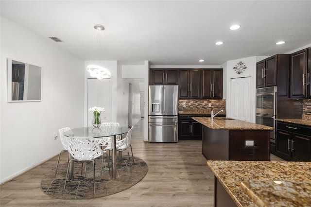 kitchen featuring a center island with sink, wood-type flooring, appliances with stainless steel finishes, hanging light fixtures, and stone counters