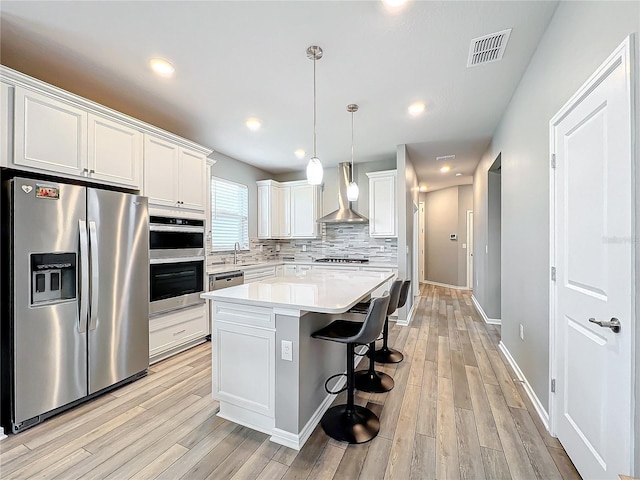 kitchen with light hardwood / wood-style floors, white cabinetry, a kitchen island, wall chimney range hood, and appliances with stainless steel finishes