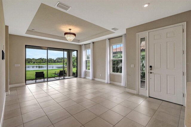 tiled foyer entrance featuring a healthy amount of sunlight, a raised ceiling, a water view, and a notable chandelier