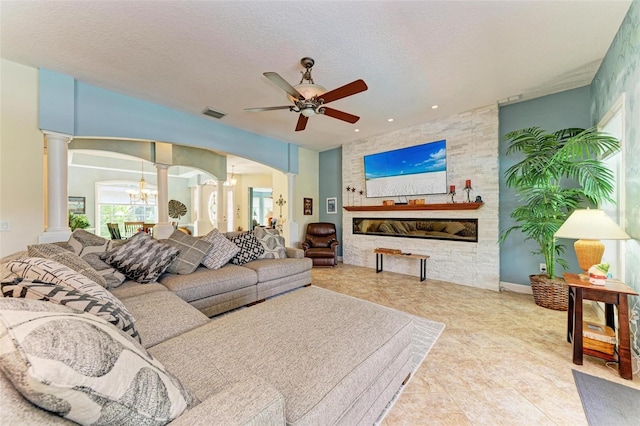 tiled living room with ceiling fan with notable chandelier, a textured ceiling, ornate columns, and a stone fireplace