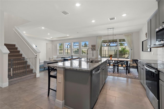 kitchen featuring stainless steel appliances, light stone counters, an island with sink, sink, and gray cabinetry