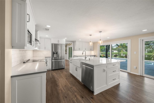 kitchen featuring white cabinetry, dark wood-type flooring, stainless steel appliances, decorative light fixtures, and a center island with sink