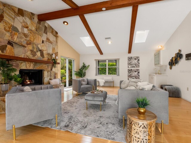 living room featuring light hardwood / wood-style flooring, a stone fireplace, high vaulted ceiling, a skylight, and beamed ceiling