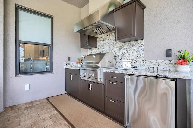 kitchen featuring light stone countertops, stainless steel refrigerator, wall chimney exhaust hood, decorative backsplash, and dark brown cabinetry