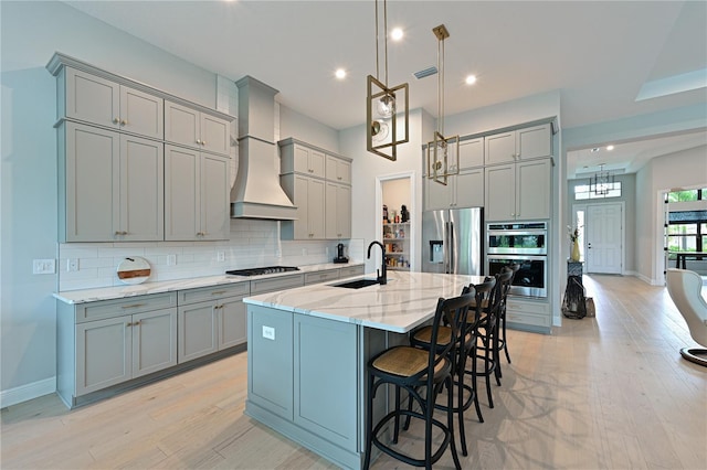 kitchen featuring light wood-type flooring, custom range hood, stainless steel appliances, sink, and an island with sink