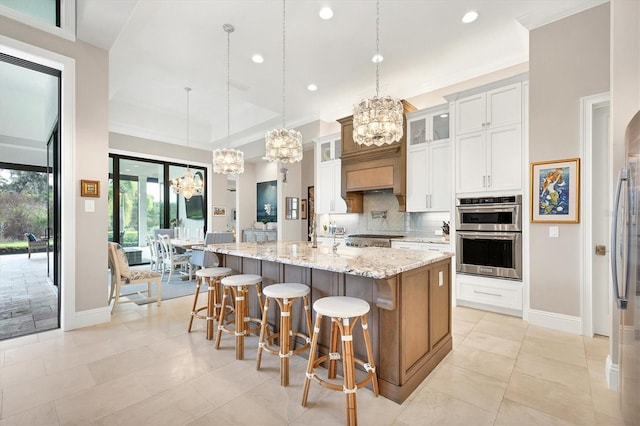 kitchen featuring stainless steel double oven, an island with sink, decorative light fixtures, white cabinets, and light stone counters