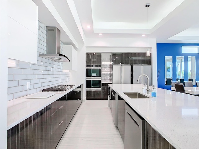 kitchen featuring appliances with stainless steel finishes, a tray ceiling, sink, light stone counters, and wall chimney range hood