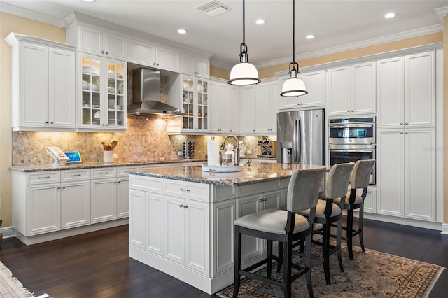 kitchen featuring wall chimney exhaust hood, stainless steel appliances, light stone countertops, and a kitchen island with sink