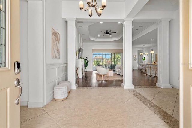 foyer entrance featuring a tray ceiling, crown molding, ornate columns, a decorative wall, and ceiling fan with notable chandelier