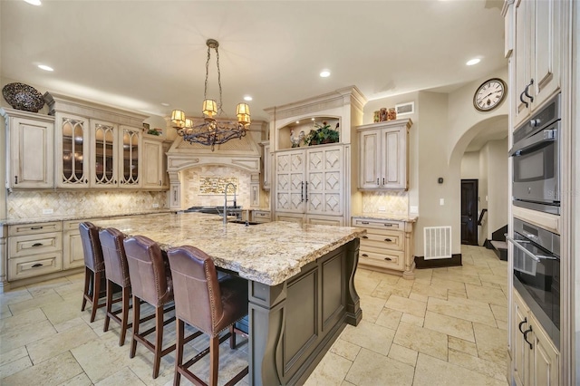 kitchen with decorative backsplash, oven, a kitchen island with sink, and light stone counters