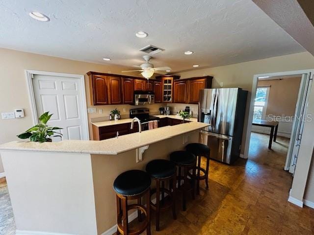 kitchen with ceiling fan, a kitchen island with sink, a breakfast bar area, a textured ceiling, and stainless steel appliances