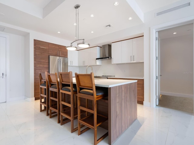 kitchen with stainless steel refrigerator, white cabinetry, wall chimney exhaust hood, decorative light fixtures, and a kitchen island with sink