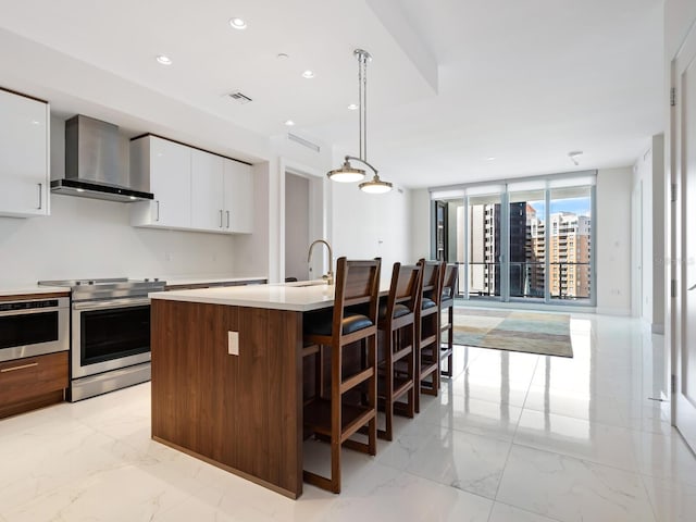 kitchen with white cabinetry, wall chimney exhaust hood, stainless steel appliances, decorative light fixtures, and a kitchen island with sink