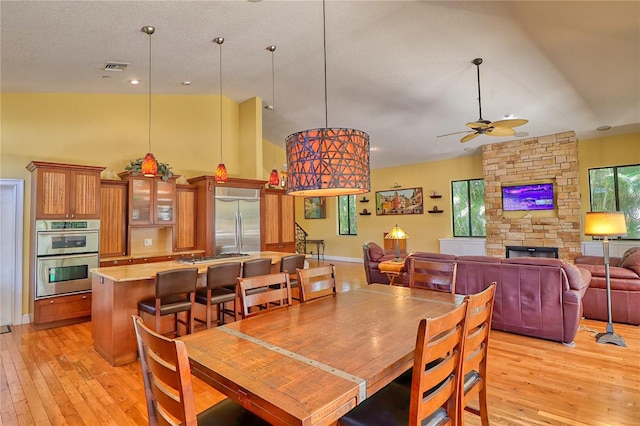 dining room with high vaulted ceiling, light wood-type flooring, ceiling fan, and a stone fireplace