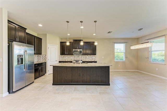 kitchen featuring decorative backsplash, a kitchen island with sink, hanging light fixtures, appliances with stainless steel finishes, and light stone counters