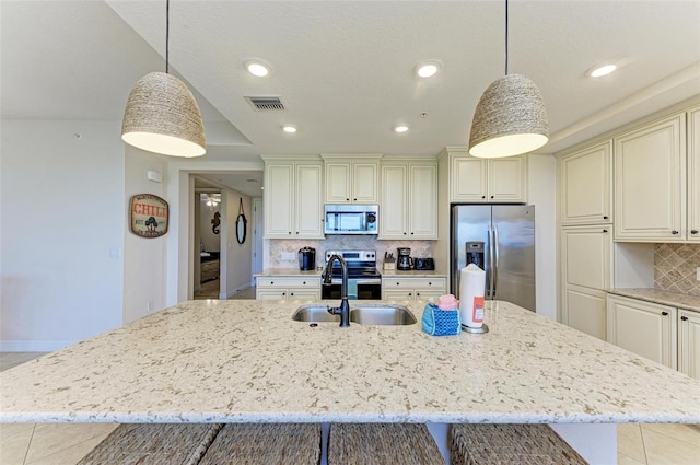 kitchen featuring decorative backsplash, stainless steel appliances, a kitchen island with sink, decorative light fixtures, and cream cabinetry