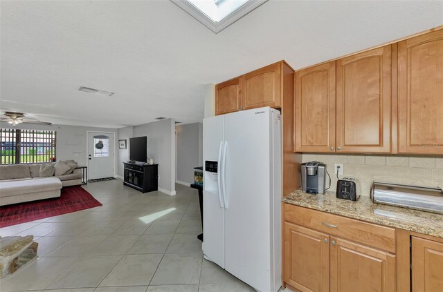 kitchen featuring white fridge with ice dispenser, a skylight, light tile patterned floors, light stone countertops, and ceiling fan