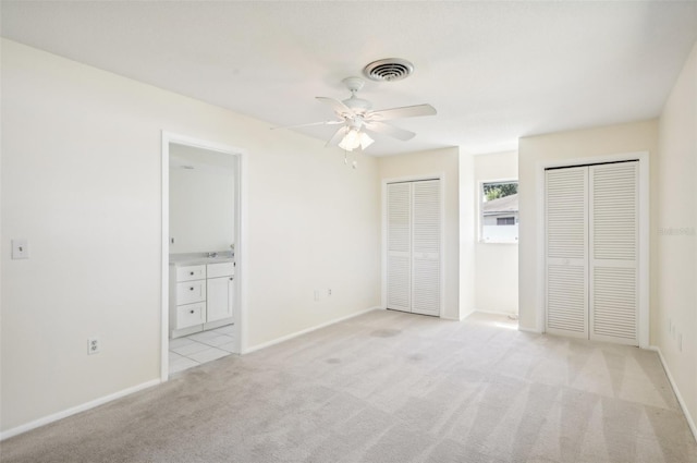 unfurnished bedroom with ceiling fan, ensuite bathroom, two closets, and light carpet