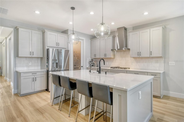 kitchen with decorative backsplash, wall chimney exhaust hood, light wood-type flooring, a center island with sink, and sink