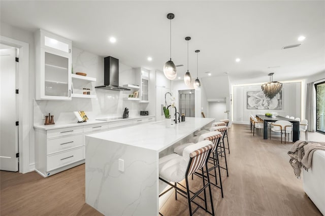 kitchen featuring white cabinetry, hanging light fixtures, wall chimney range hood, a kitchen island with sink, and light wood-type flooring