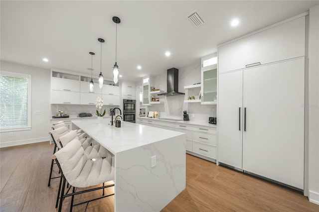 kitchen featuring white cabinetry, wall chimney exhaust hood, light hardwood / wood-style flooring, a large island with sink, and a kitchen bar