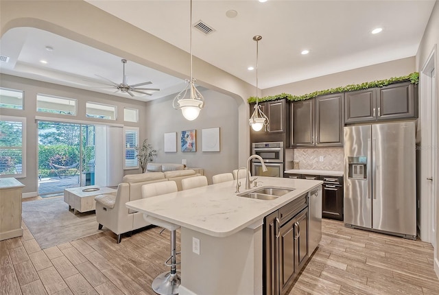 kitchen featuring stainless steel appliances, a center island with sink, sink, hanging light fixtures, and light hardwood / wood-style flooring