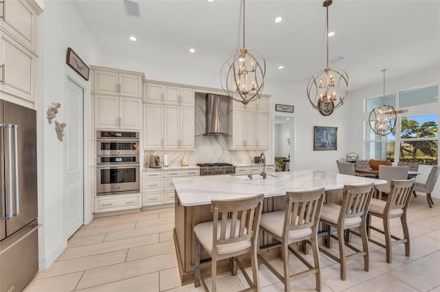 kitchen featuring hanging light fixtures, a kitchen island with sink, appliances with stainless steel finishes, cream cabinetry, and wall chimney exhaust hood