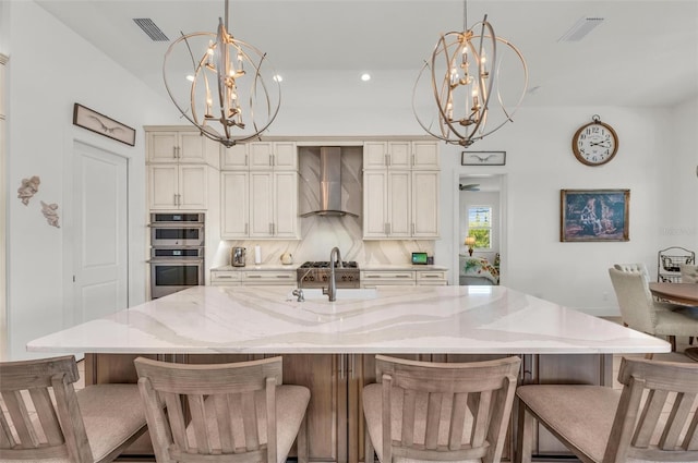 kitchen with cream cabinetry, backsplash, wall chimney range hood, hanging light fixtures, and double oven