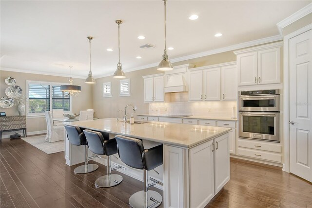 kitchen featuring stainless steel double oven, hanging light fixtures, a kitchen island with sink, and dark hardwood / wood-style flooring