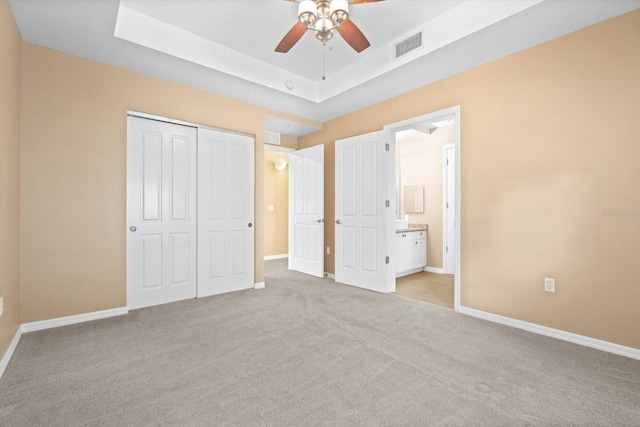 unfurnished bedroom featuring ensuite bath, a closet, light colored carpet, a tray ceiling, and ceiling fan