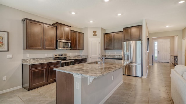 kitchen with dark brown cabinetry, light stone countertops, a kitchen island with sink, light tile patterned flooring, and appliances with stainless steel finishes