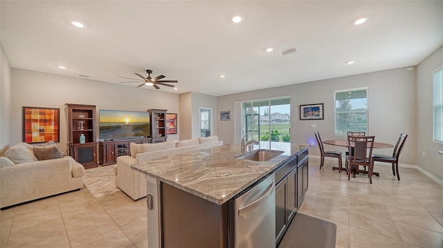 kitchen featuring a center island with sink, sink, stainless steel dishwasher, ceiling fan, and light stone countertops