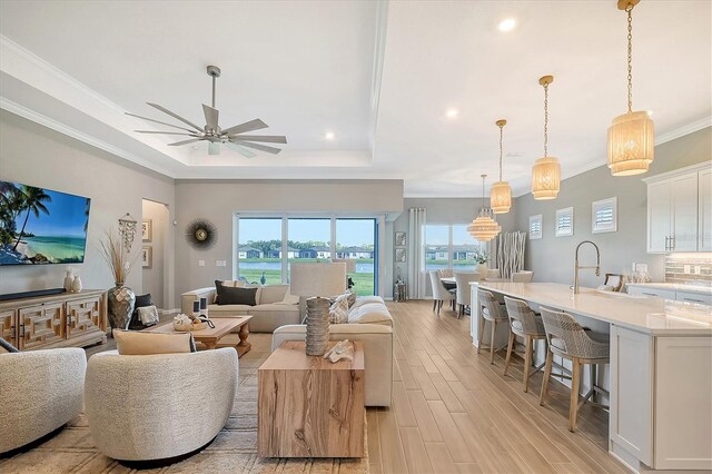 living room featuring ceiling fan, ornamental molding, sink, a tray ceiling, and light hardwood / wood-style floors