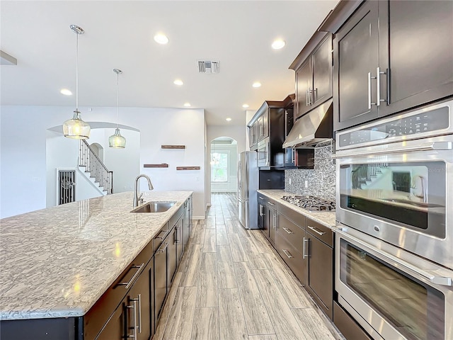 kitchen with backsplash, a kitchen island with sink, sink, decorative light fixtures, and stainless steel appliances