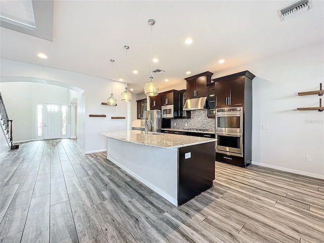 kitchen featuring a kitchen island with sink, decorative backsplash, light stone countertops, appliances with stainless steel finishes, and decorative light fixtures