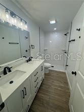bathroom featuring a sink, toilet, double vanity, wood finished floors, and tiled shower
