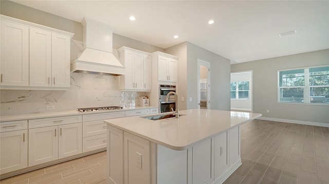 kitchen with a kitchen island with sink, custom range hood, sink, white cabinets, and appliances with stainless steel finishes