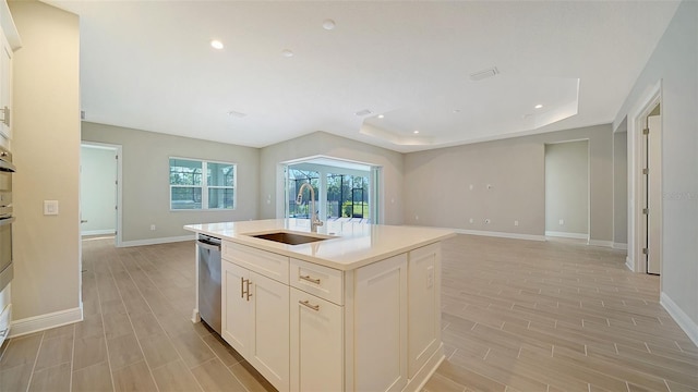 kitchen with light wood-type flooring, a kitchen island with sink, stainless steel dishwasher, sink, and a tray ceiling