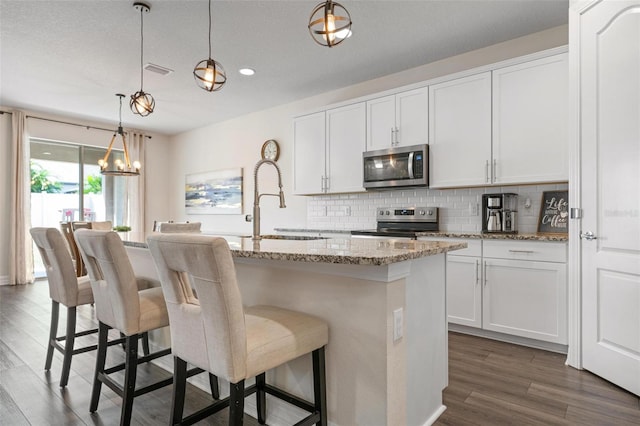 kitchen with light stone counters, white cabinets, decorative light fixtures, a kitchen island with sink, and appliances with stainless steel finishes