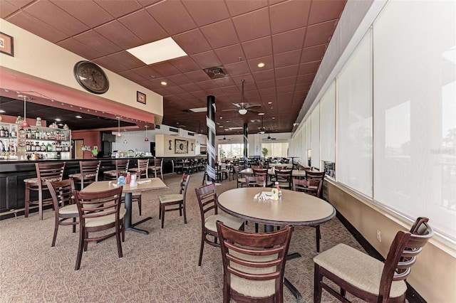 carpeted dining area with a drop ceiling, ceiling fan, and indoor bar