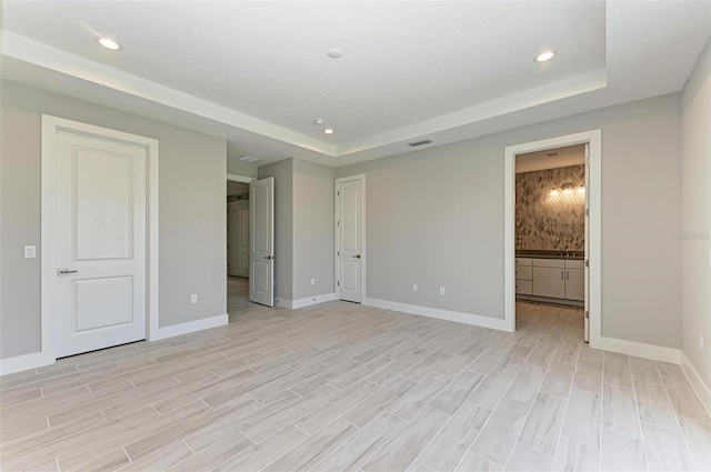 unfurnished bedroom with light wood-type flooring, a textured ceiling, connected bathroom, and a tray ceiling