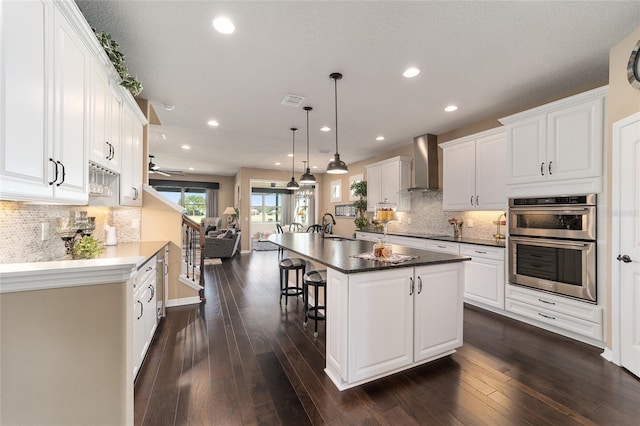 kitchen featuring pendant lighting, wall chimney exhaust hood, an island with sink, dark hardwood / wood-style flooring, and white cabinetry