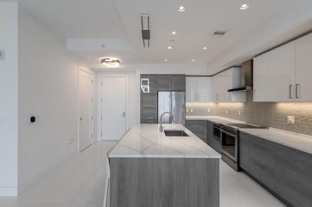 kitchen featuring sink, an island with sink, white cabinets, wall chimney range hood, and appliances with stainless steel finishes